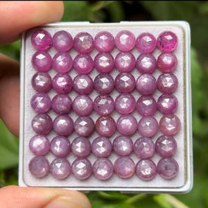 May include: A collection of faceted, round, pink gemstones arranged in a white tray. The gemstones have a polished, reflective surface and are of varying shades of pink. The tray is held by a hand.