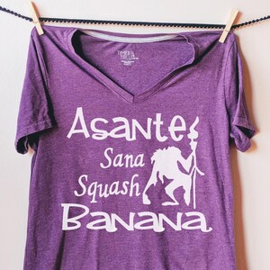 May include: Purple V-neck t-shirt with a white graphic of a cartoon character holding a torch and the text "Asante Sana Squash Banana".