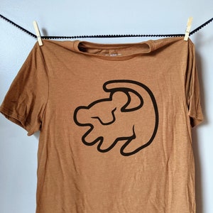 May include: A brown t-shirt with a black outline of Simba's paw print from the Lion King movie.