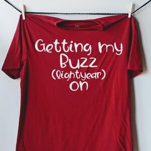 May include: A red t-shirt with the phrase "Getting my Buzz (lightyear) on" in white lettering. The shirt is hanging from a clothesline with a wooden clothespin.