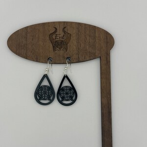 May include: Black teardrop-shaped earrings with a silver hook and a black geometric design inside each teardrop. The earrings are hanging from a wooden display stand with the text "Evil Queen Designs" engraved on it.