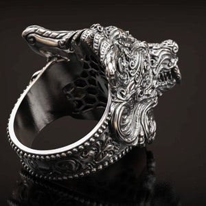 May include: A silver-colored ring with an ornate design featuring a detailed dragon or mythical creature head. The ring has a textured surface and intricate patterns, with a dark background.