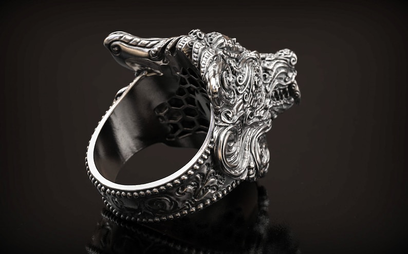May include: A silver-colored ring with an ornate design featuring a detailed dragon or mythical creature head. The ring has a textured surface and intricate patterns, with a dark background.