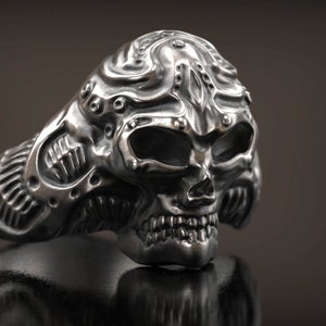 Könnte beinhalten: Ein detaillierter silberner Totenkopfring mit aufwendigen, strukturierten Details. Der Ring hat ein Totenkopfdesign mit offenen Augenhöhlen und einem detaillierten Kiefer. Das Design des Rings umfasst Nieten und eine strukturierte Oberfläche.