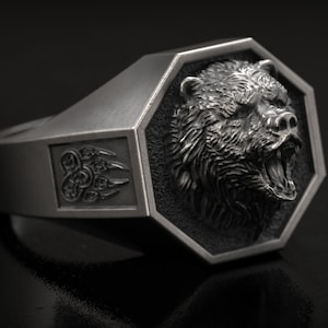 May include: A silver ring featuring a detailed bear head within a geometric frame. The ring has a rectangular design with a bear claw emblem on the side. The bear's mouth is open, revealing its teeth.