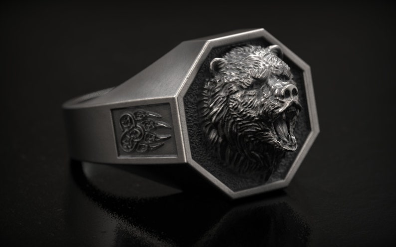 May include: A silver ring featuring a detailed bear head within a geometric frame. The ring has a rectangular design with a bear claw emblem on the side. The bear's mouth is open, revealing its teeth.