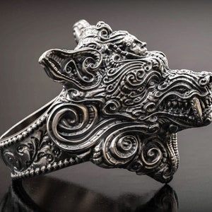 May include: A detailed silver ring featuring a stylized wolf head design. The ring showcases intricate scrollwork and a fierce expression, with visible teeth. The ring is a statement piece, perfect for those who appreciate unique jewelry.