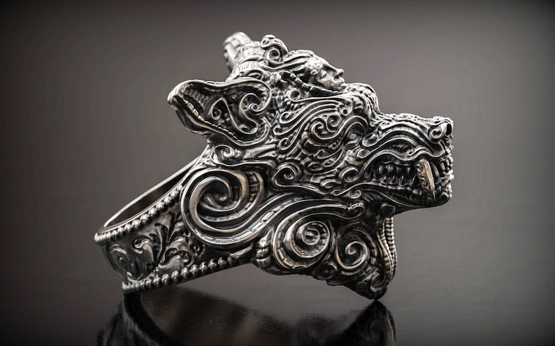 May include: A detailed silver ring featuring a stylized wolf head design. The ring showcases intricate scrollwork and a fierce expression, with visible teeth. The ring is a statement piece, perfect for those who appreciate unique jewelry.