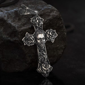 Könnte beinhalten: Silberne Anhänger Halskette mit detailliertem Kreuz-Design. Das Kreuz ist mit Rosen und einer zentralen Figur mit Bandana verziert. Der Anhänger hängt an einem schwarzen Band vor dunklem, strukturiertem Hintergrund.