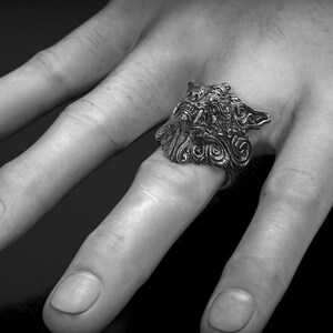 May include: A detailed silver ring featuring a wolf's head design. The ring is worn on a finger, showcasing intricate carvings and a textured finish. The image is in black and white, highlighting the ring's details.