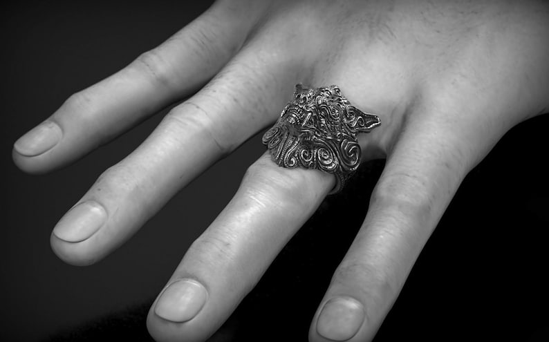 May include: A detailed silver ring featuring a wolf's head design. The ring is worn on a finger, showcasing intricate carvings and a textured finish. The image is in black and white, highlighting the ring's details.