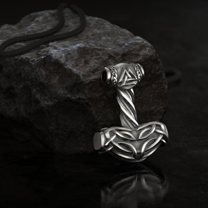 Könnte beinhalten: Eine silberfarbene Halskette mit Anhänger, die Thors Hammer, Mjolnir, darstellt. Der Hammer hat aufwendige Knotenarbeiten und einen gedrehten Griff, mit einer schwarzen Kordel. Der Anhänger liegt auf einem dunklen, strukturierten Stein.