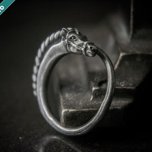 May include: A silver-colored ring with a horse head design. The horse head is detailed with a braided mane and a bit in its mouth. The ring is set against a dark background, highlighting the intricate details of the jewelry.