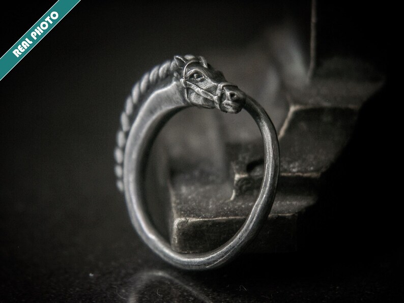 May include: A silver-colored ring with a horse head design. The horse head is detailed with a braided mane and a bit in its mouth. The ring is set against a dark background, highlighting the intricate details of the jewelry.