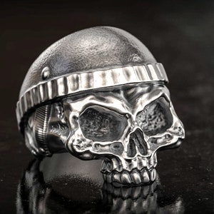 Könnte beinhalten: Ein silberner Totenkopfring mit Helmdesign. Der Ring zeigt ein detailliertes Totenkopfgesicht mit sichtbaren Zähnen und Augenhöhlen. Der Helm hat eine genietete Textur und ein Band um den Kopf. Der Ring besteht aus glänzendem Metall.