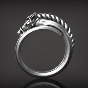 May include: A silver-colored ring featuring a horse head design. The horse head is detailed with a bridle and a braided mane that curves around the ring's band. The ring is set against a dark background.