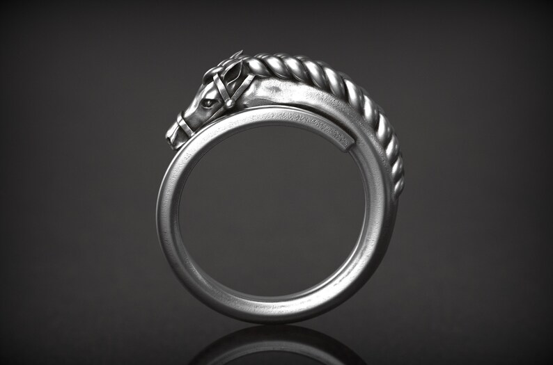 May include: A silver-colored ring featuring a horse head design. The horse head is detailed with a bridle and a braided mane that curves around the ring's band. The ring is set against a dark background.