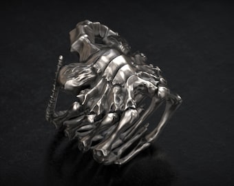 May include: A silver-colored ring with a detailed skeletal design. The ring features a complex arrangement of bones, ribs, and a spine-like structure, creating a gothic or fantasy aesthetic. The ring is displayed on a dark, textured surface.