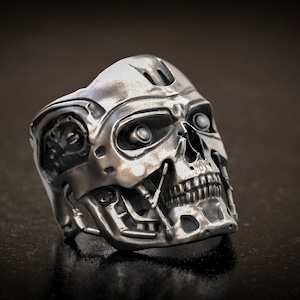 May include: A silver-colored ring shaped like a robotic skull. The ring features detailed mechanical elements, including eye sockets, teeth, and head components. The ring is designed with a dark background.