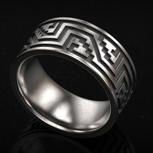 May include: A wide, silver-colored ring with a geometric pattern. The ring features a repeating design of angular shapes and lines around the band. The ring is displayed against a dark background, highlighting its metallic sheen.