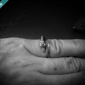 May include: A silver ring with a braided design and a small, decorative element. The ring is worn on a finger, showcasing its intricate details and craftsmanship. The image is in black and white.