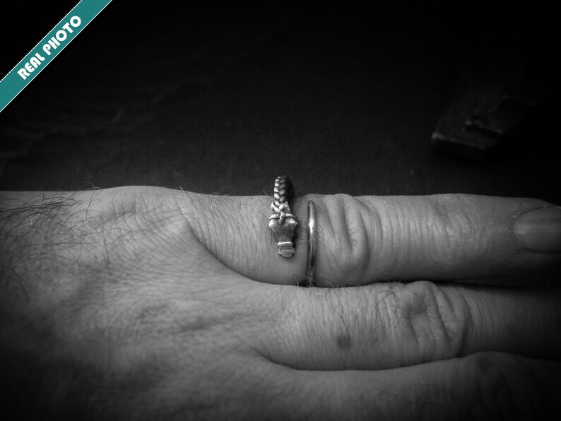 May include: A silver ring with a braided design and a small, decorative element. The ring is worn on a finger, showcasing its intricate details and craftsmanship. The image is in black and white.