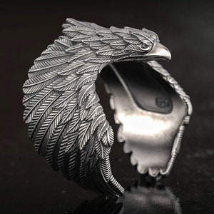 Könnte beinhalten: Ein silberfarbenes Armreif-Armband mit einem Adler-Design. Das Armband ist mit detaillierten Federn und einem Adlerkopf geformt, was ein auffälliges Accessoire ergibt. Das Armband ist unten offen.