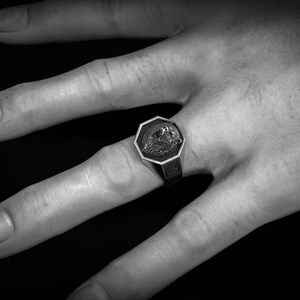 May include: A silver-toned signet ring with an octagonal face featuring a detailed relief design. The ring is worn on a hand against a dark background. The ring's design is centered within the octagonal frame.