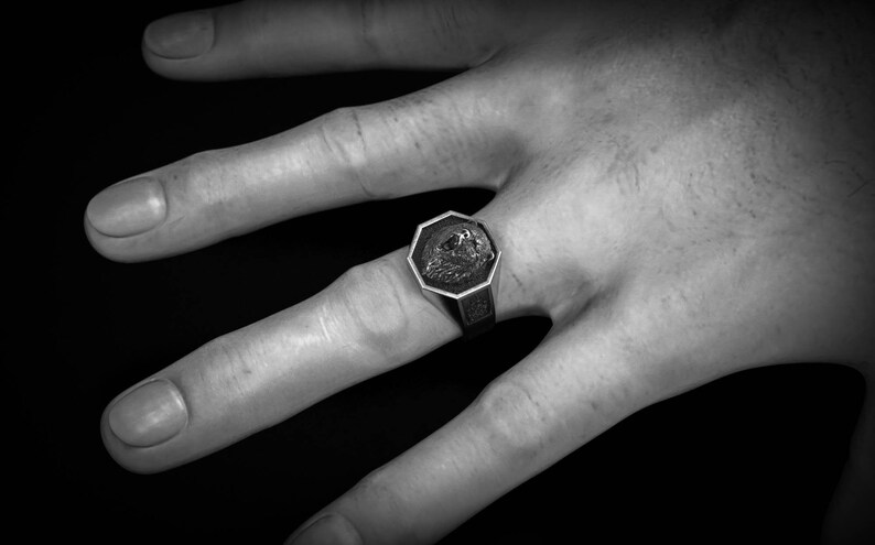 May include: A silver-toned signet ring with an octagonal face featuring a detailed relief design. The ring is worn on a hand against a dark background. The ring's design is centered within the octagonal frame.