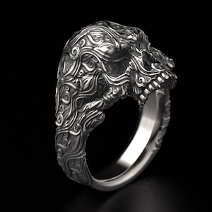Könnte beinhalten: Ein detaillierter Silberring mit einem Totenkopf-Design. Der Totenkopf ist aufwendig mit wirbelnden Mustern und Texturen geschnitzt. Der Ring ist vor einem schwarzen Hintergrund platziert, der den Glanz des Metalls hervorhebt.