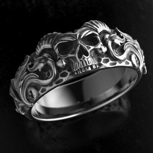 Könnte beinhalten: Ein silberfarbener Ring mit einem Totenkopf-Design. Der Ring zeigt einen detaillierten Totenkopf in der Mitte, umgeben von kunstvollen, wirbelnden Mustern. Der Ring ist vor einem dunklen Hintergrund platziert, der seine komplizierten Details hervorhebt.