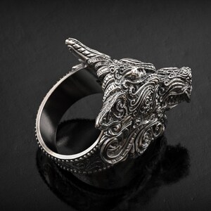 May include: A silver-colored ring featuring a detailed dragon head design. The dragon's head is intricately carved with scales, horns, and facial features. The ring has a wide band and is displayed against a dark background.