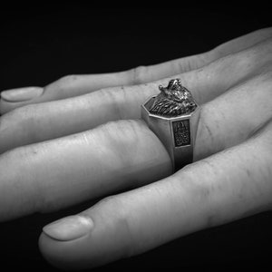 May include: A silver ring with a wolf head design. The ring has an octagonal face with a detailed wolf head and a decorative emblem on the band. The ring is shown on a hand against a dark background.