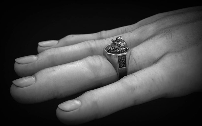 May include: A silver ring with a wolf head design. The ring has an octagonal face with a detailed wolf head and a decorative emblem on the band. The ring is shown on a hand against a dark background.