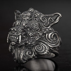 May include: A detailed silver ring featuring a stylized wolf's head with intricate carvings. The ring showcases sharp teeth and a textured surface, set against a dark background. The design is bold and eye-catching.