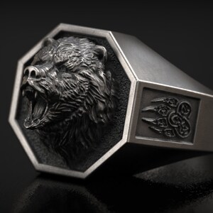 May include: A silver-colored ring with a detailed bear head design. The bear's mouth is open, revealing its teeth. The ring has an octagonal face and a side with a bear claw design. A bold, statement piece.