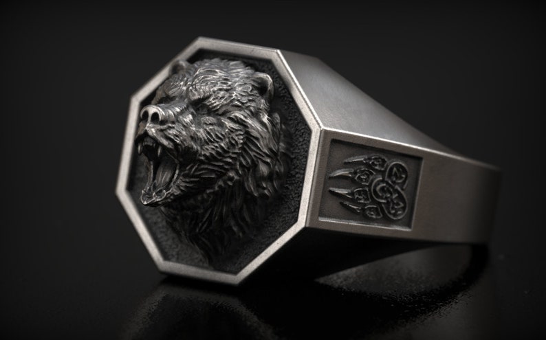 May include: A silver-colored ring with a detailed bear head design. The bear's mouth is open, revealing its teeth. The ring has an octagonal face and a side with a bear claw design. A bold, statement piece.