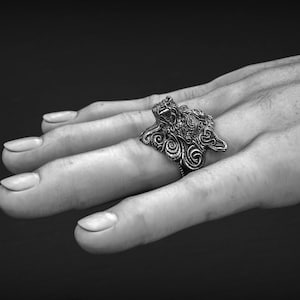 May include: A detailed silver ring with an ornate, swirling design is displayed. The ring is worn on a finger, showcasing its intricate patterns and textures. The ring's design features a central figure with surrounding swirls.