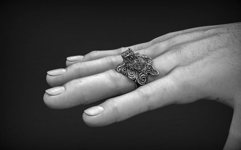 May include: A detailed silver ring with an ornate, swirling design is displayed. The ring is worn on a finger, showcasing its intricate patterns and textures. The ring's design features a central figure with surrounding swirls.
