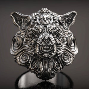 May include: A detailed silver ring featuring a stylized wolf's head design. The ring showcases intricate carvings, including a central face with bared teeth and swirling patterns. The ring is displayed against a dark background.