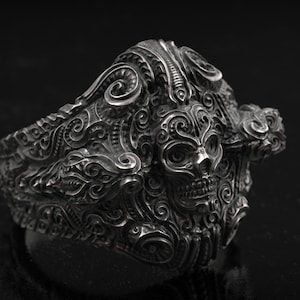 Puede incluir: Un anillo de plata detallado con un diseño de calavera. El anillo tiene patrones intrincados y un acabado oscuro y envejecido. La calavera es el punto focal, rodeada de detalles ornamentados y arremolinados. El anillo es una pieza audaz.