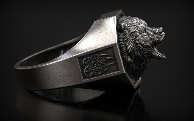 May include: A silver-colored ring featuring a detailed bear head design. The ring has a rectangular accent with a claw symbol. The bear's mouth is open, and the ring is set against a dark background.