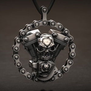 Può includere: Ciondolo color argento con un design a motore di moto con un teschio al centro. Il motore è circondato da una catena, sospeso da un cordino nero. Il ciondolo ha un aspetto metallico dettagliato.