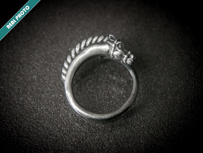 May include: A silver-toned ring featuring a horse head design. The horse head is detailed with a bridle and mane, and the ring band has a rope-like texture. The ring is set against a dark background.