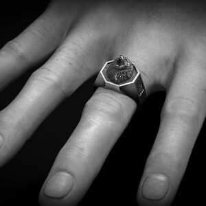 May include: A silver-toned ring featuring a detailed wolf head design. The ring has an octagonal face and is worn on a finger. The image is in black and white, highlighting the ring's intricate details and the texture of the metal.