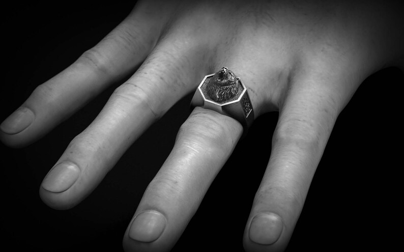 May include: A silver-toned ring featuring a detailed wolf head design. The ring has an octagonal face and is worn on a finger. The image is in black and white, highlighting the ring's intricate details and the texture of the metal.