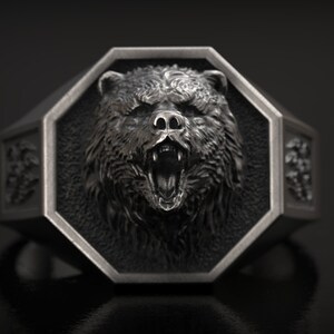 May include: A silver-colored ring with a detailed bear head design. The bear's face is centered on an octagonal face, with an open mouth and visible teeth. The ring has decorative side panels and a dark background.