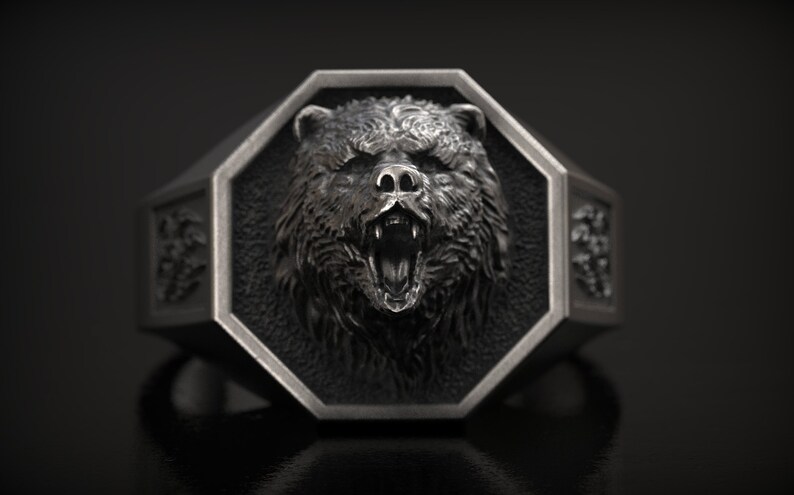 May include: A silver-colored ring with a detailed bear head design. The bear's face is centered on an octagonal face, with an open mouth and visible teeth. The ring has decorative side panels and a dark background.