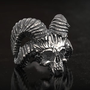 Könnte beinhalten: Ein silberfarbener Ring mit einem detaillierten Totenkopf-Design und großen, gebogenen Hörnern. Der Ring hat ein gotisches oder Fantasy-Thema mit aufwendigen Details an Schädel und Hörnern. Der Ring ist vor einem dunklen Hintergrund platziert.