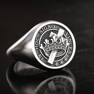 Könnte beinhalten: Silberner Siegelring mit detailliertem Emblem. Er zeigt eine Krone, ein Kreuz und die Inschrift „IN HOC SIGNO VINCES“. Der Ring hat eine polierte Oberfläche und ein klassisches, abgerundetes Design. Ein Symbol des Glaubens und des Erbes.